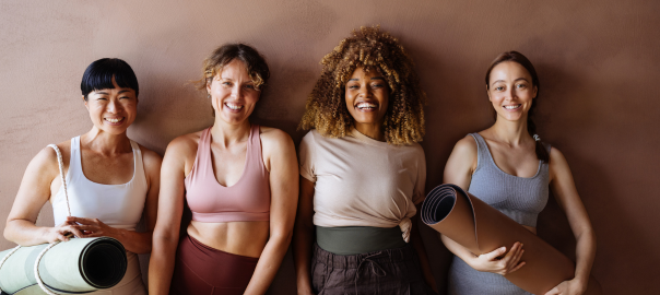 Four mid-life women stood in a row wearing yoga attire. The two women on the end are holding yoga mats. The background is a pale, beige pink.