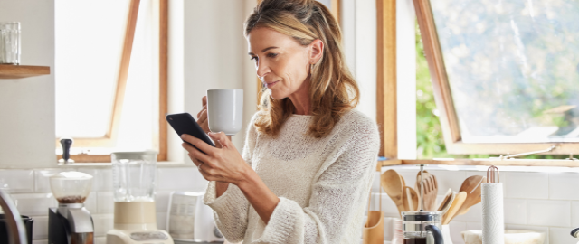 Stylish 50 something woman in her kitchen holding a mug and phone researching whether antioxidants can help the immune system.