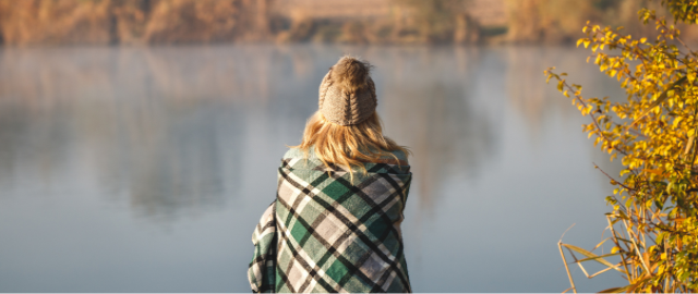 Women with her back to the camera, sat next to a lake in autumn, wrapped in a tartan blanket and wearing a woolly hat.