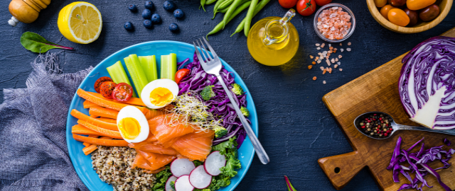 Flatlay of colourful, healthy food: a bowl of salmon, quinoa and salad in a blue bowl on a dark blue table with red cabbage, blueberries, olive oil and laid on the table.