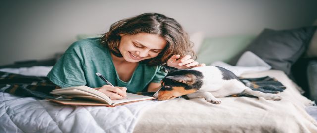 Woman in aturquoise blue top journalling while lay on a cream bed, with a dog next to her.