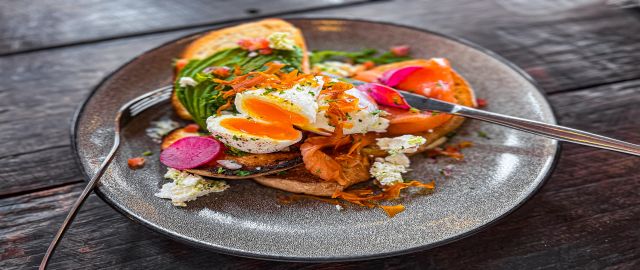 Horizontal high angle closeup photo of free range poached eggs on toasted buttered sourdough bread with smoked salmon, pickled radish slices, sliced avocado, feta cheese, sweet potato crisps and salad leaves on a ceramic plate on a rustic wooden table in 
