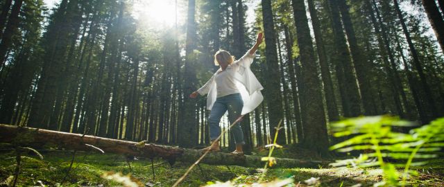 Playful mid-life woman enjoying nature. She is barefoot, balancing with outstretched arms on a fallen log in the forest.