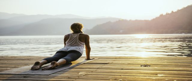 Woman doing yoga by a lake at sunrise. She is in upward dog pose and wears a vest and leggings. Yoga can help to balance stress and cortisol levels.