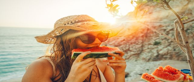 Women eating and enjoying hydrating watermelon on a beach. She wears a subhat and sunglasses and the sea is behind her.