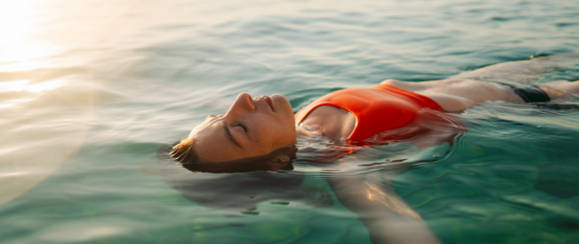 Mid-life women wearing a red swimwear top and black bottoms floats in the sea at sunset to cool off.