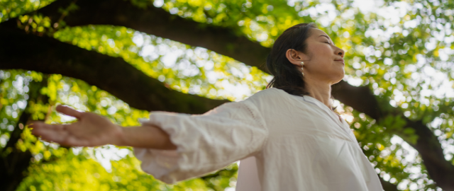Women stood underneath a tree in the woods with arms outstretched as she does a breathing practice.