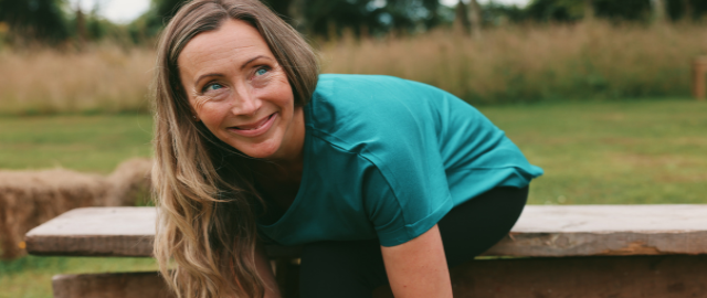 Smiling mid-life woman with long hair sits on an outdoor wooden bench by a lake to tie her shoe laces before going for a run. She wears a teal t-shirt and black leggings.
