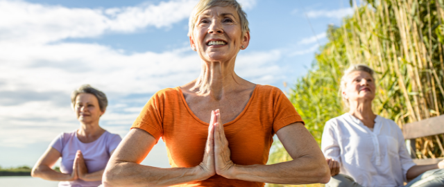 Stepping into spring: three mature women sat cross legged outdoors alongside a lake practising yoga.