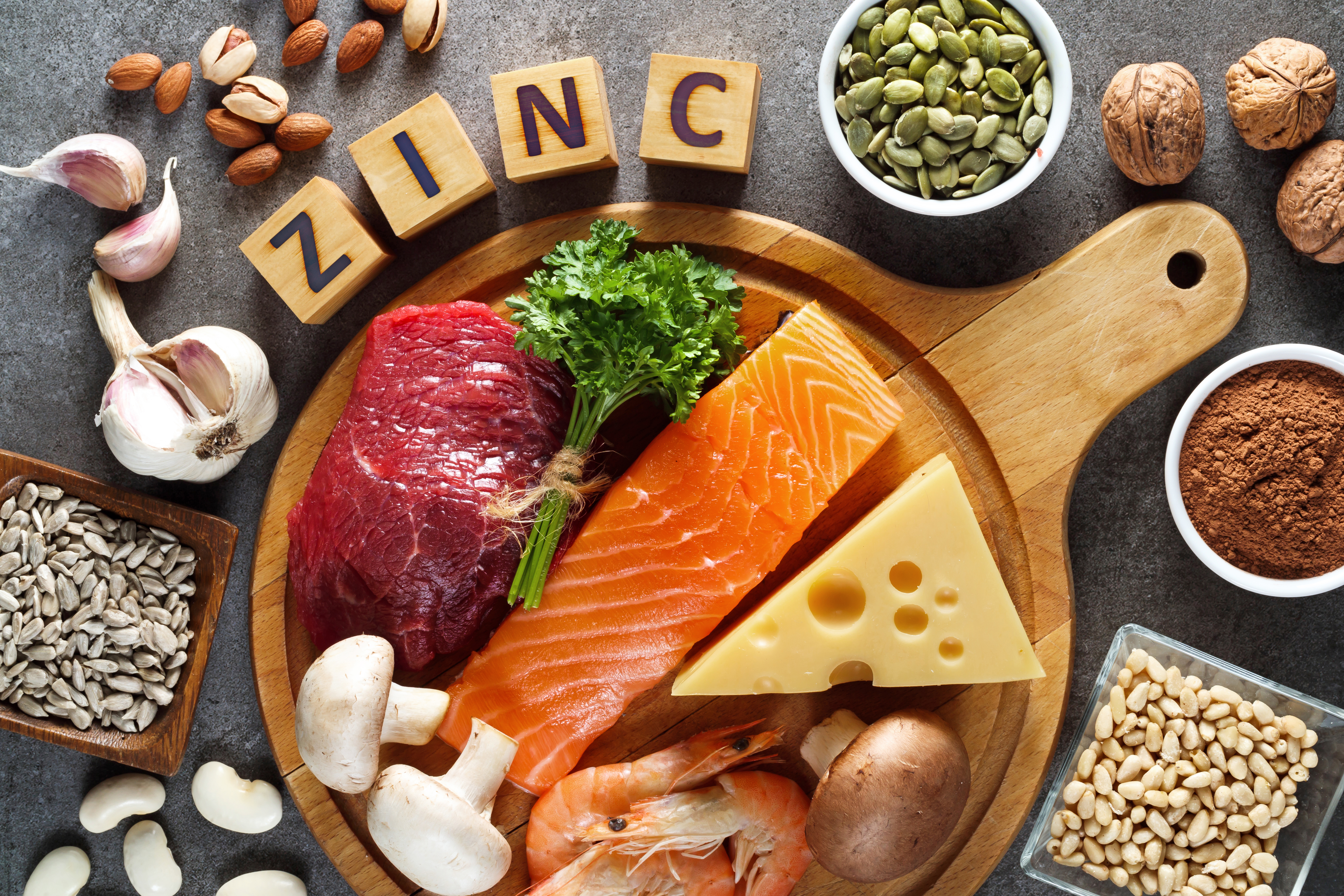 A flatlay of some of the best food sources of zinc on a round wooden serving board against a grey background. Zinc food sources of animal origin products such as meat, fish, cheese and eggs are laid out alongside vegan and plant-based sources such as pump