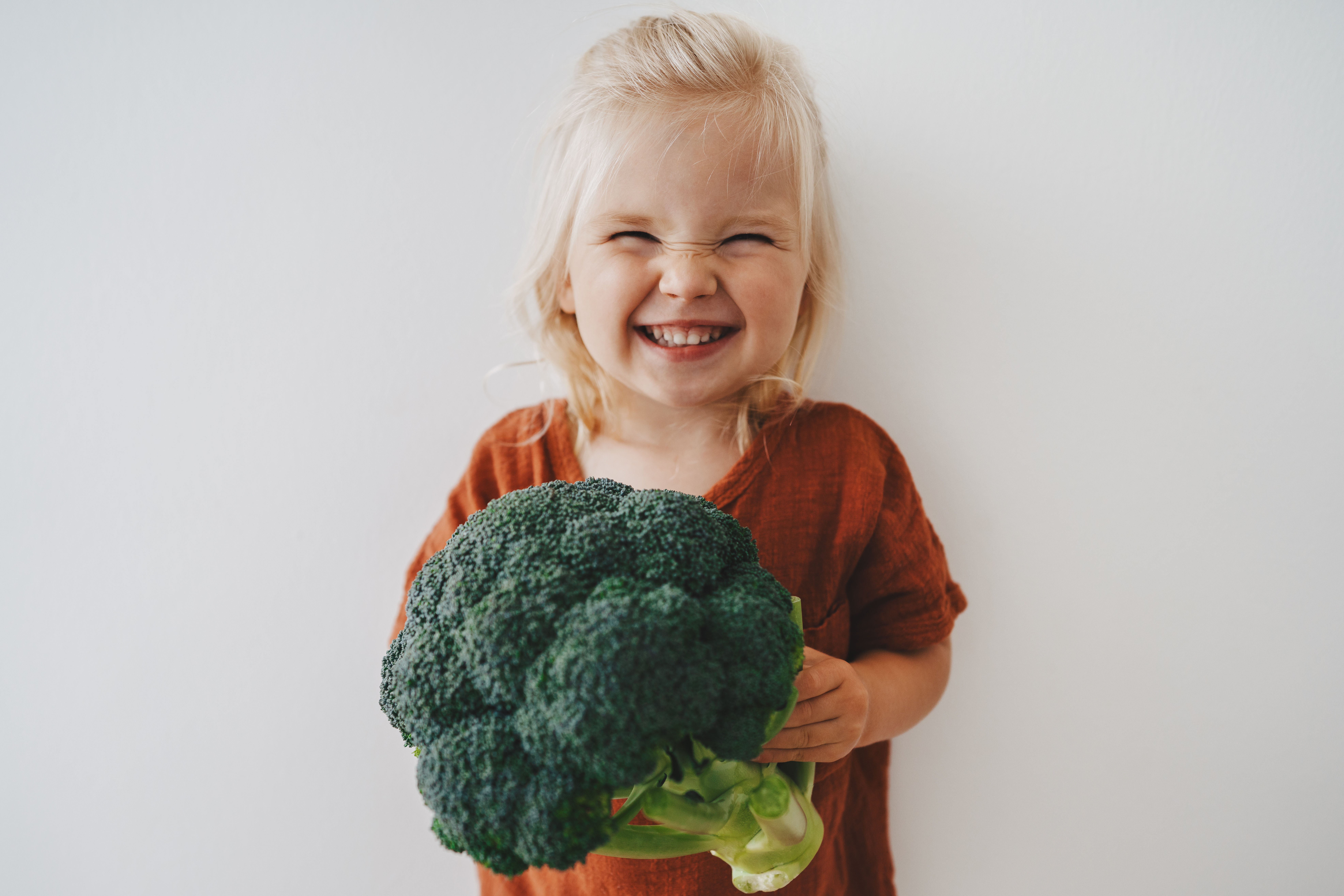 Healthy eating for children: blonde haired toddler girl with a cheeky grin holding broccoli.