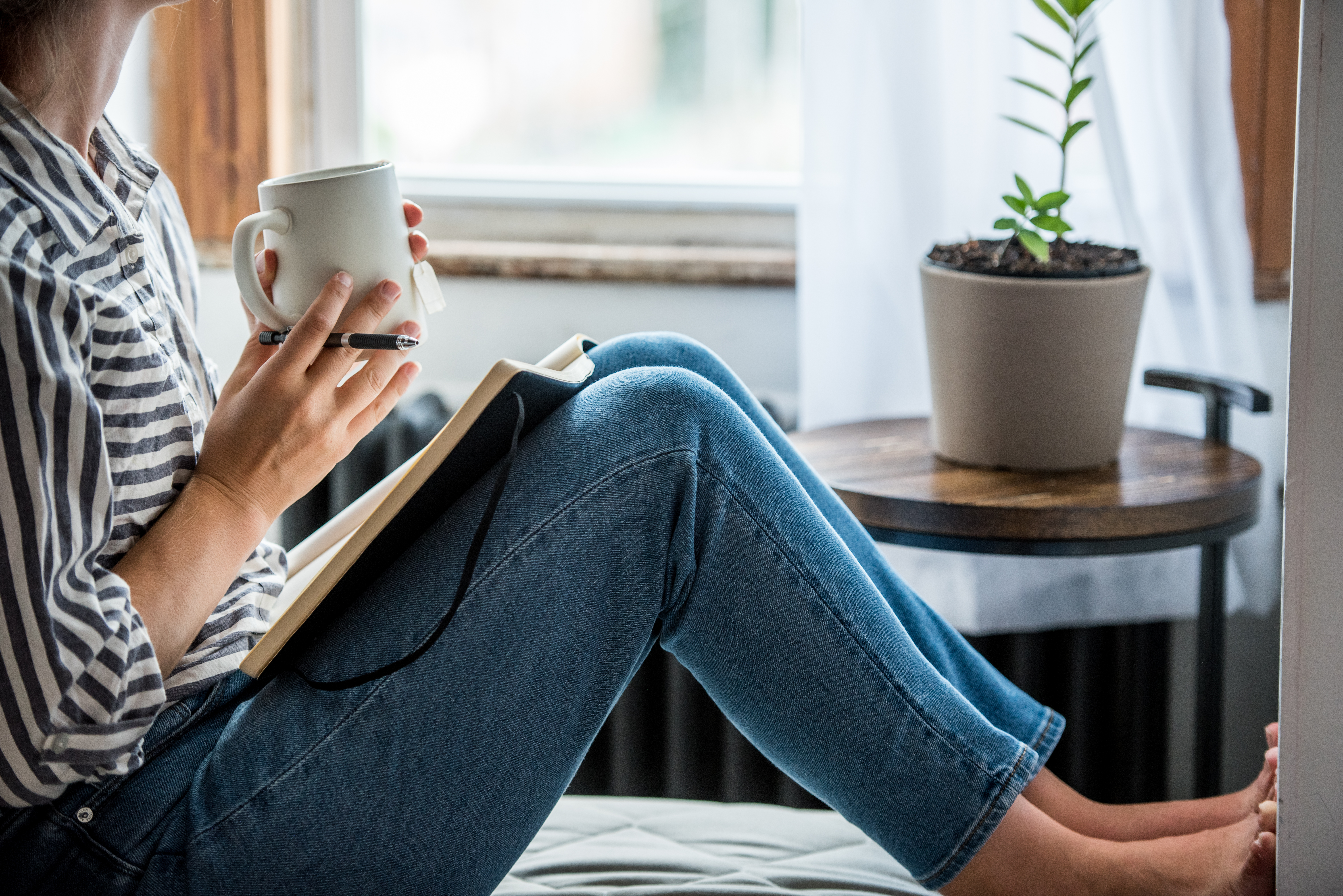 Young woman sat holding a mug whilst journalling. Journalling may help when trying to build and reinforce healthful habits.