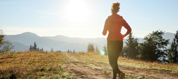 Rear view of a mid life woman running on a mountain range. She wears a red jacket and black running tights, the sun is shining and she is looking out across the top of the mountain range.