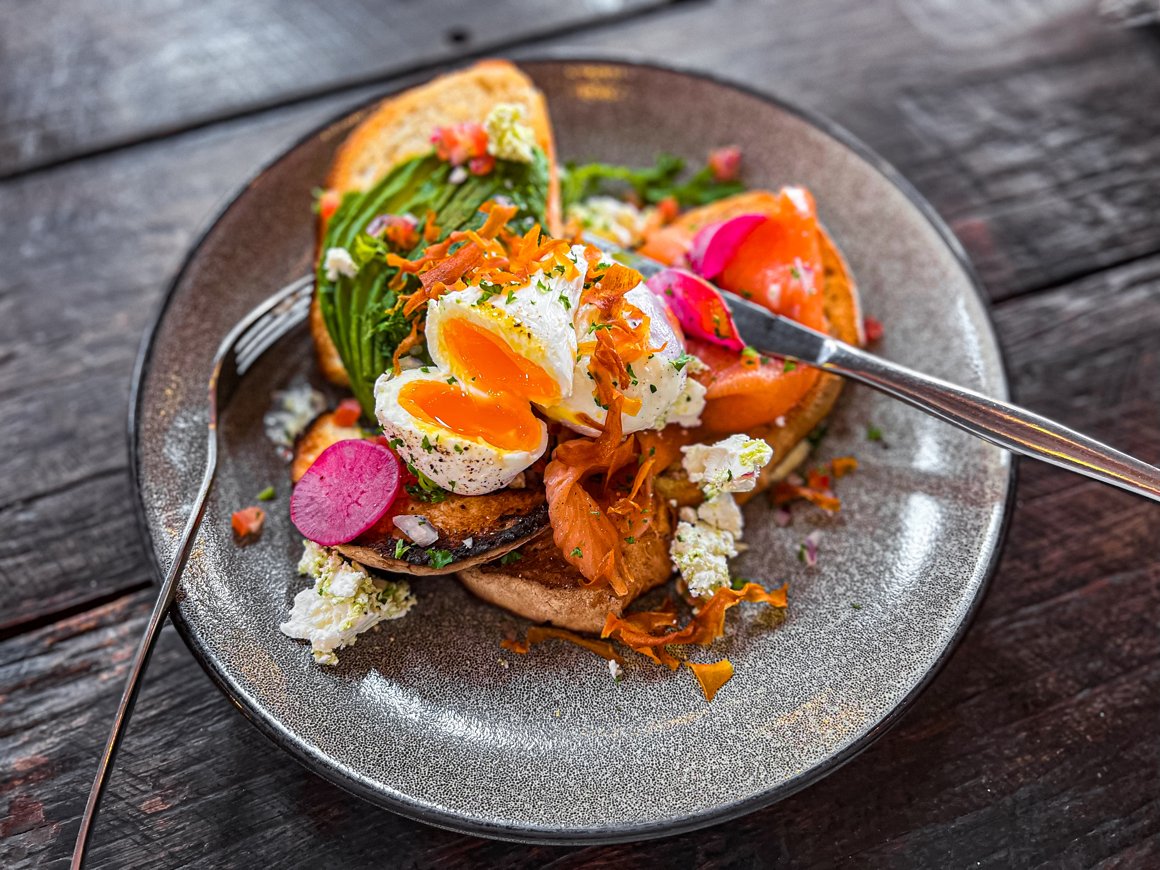 Horizontal high angle closeup photo of free range poached eggs on toasted buttered sourdough bread with smoked salmon, pickled radish slices, sliced avocado, feta cheese, sweet potato crisps and salad leaves on a ceramic plate on a rustic wooden table in